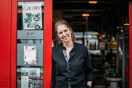 Eine Frau in schwarzer Arbeitskleidung steht im Eingang eines Lokals mit rotem Türrahmen und blickt in die Kamera.