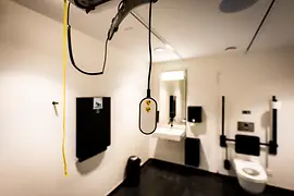 Inside an accessible restroom featuring a ceiling-mounted lift system with a hanging remote control, a sink and mirror, and support rails beside the toilet.