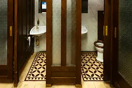 An interior view of a historic public restroom with dark wooden doors, frosted glass panels, patterned tile flooring, and vintage sinks with marble walls.