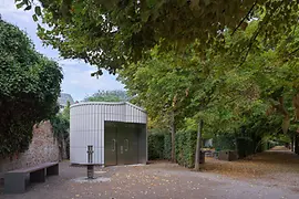 A modern public restroom building with metallic paneling, set along a tree-lined path in Augarten. Leaves cover parts of the ground, and benches and hedges frame the quiet park setting.