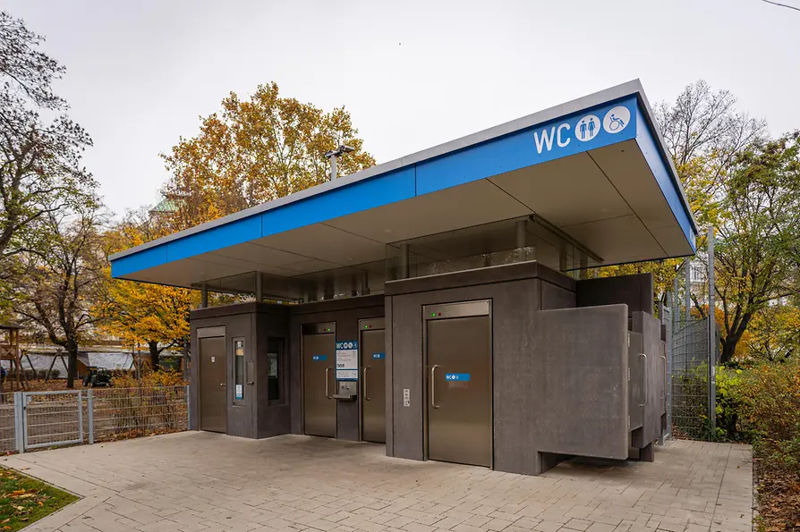 A modern public restroom facility with a blue roofline, dark gray walls, stainless-steel doors, and accessible-entry signage, surrounded by autumn foliage.