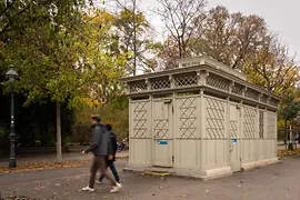 A historic light-gray public restroom pavilion with decorative lattice panels and stained-glass details, set among autumn trees as pedestrians walk by.