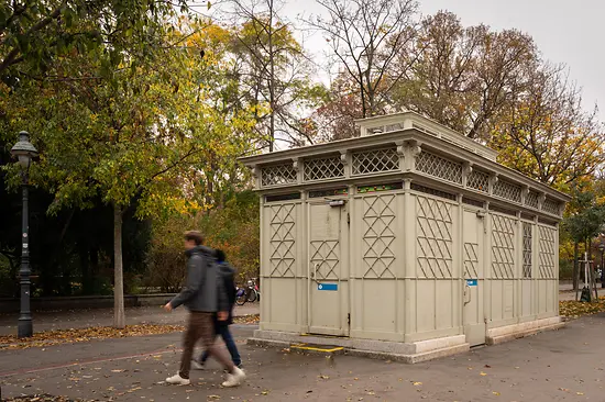 A historic light-gray public restroom pavilion with decorative lattice panels and stained-glass details, set among autumn trees as pedestrians walk by.