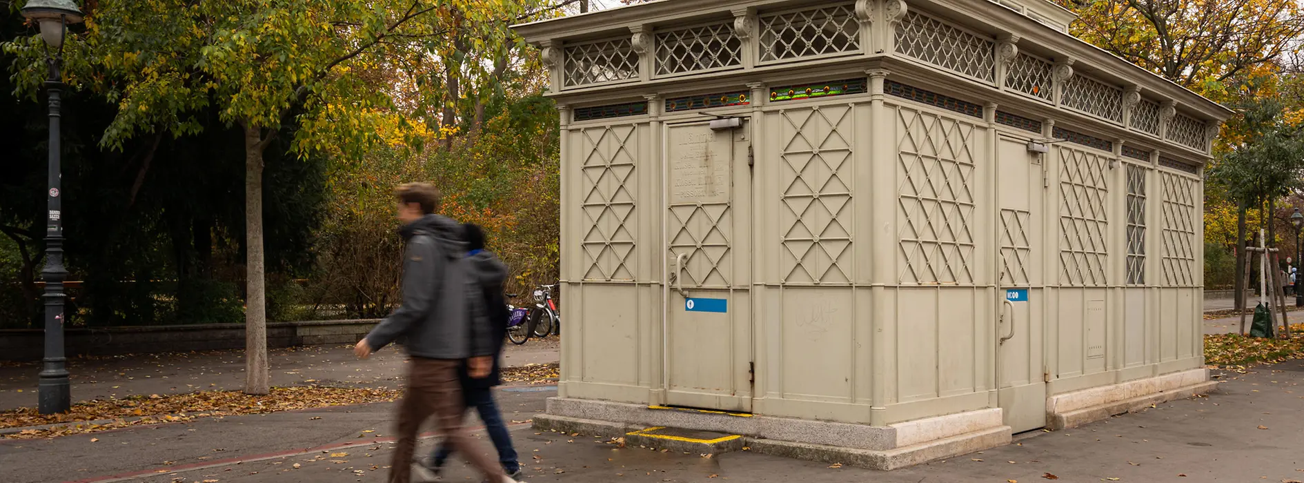 A historic light-gray public restroom pavilion with decorative lattice panels and stained-glass details, set among autumn trees as pedestrians walk by.