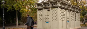 Egy történelmi, világosszürke WC-pavilon díszes rácselemekkel és színes üvegrészletekkel, őszi fákkal körülvéve, miközben gyalogosok haladnak el mellette.