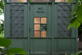 A green historic men’s restroom kiosk with decorative metalwork and frosted windows, partially hidden behind lush leafy branches in a park setting.