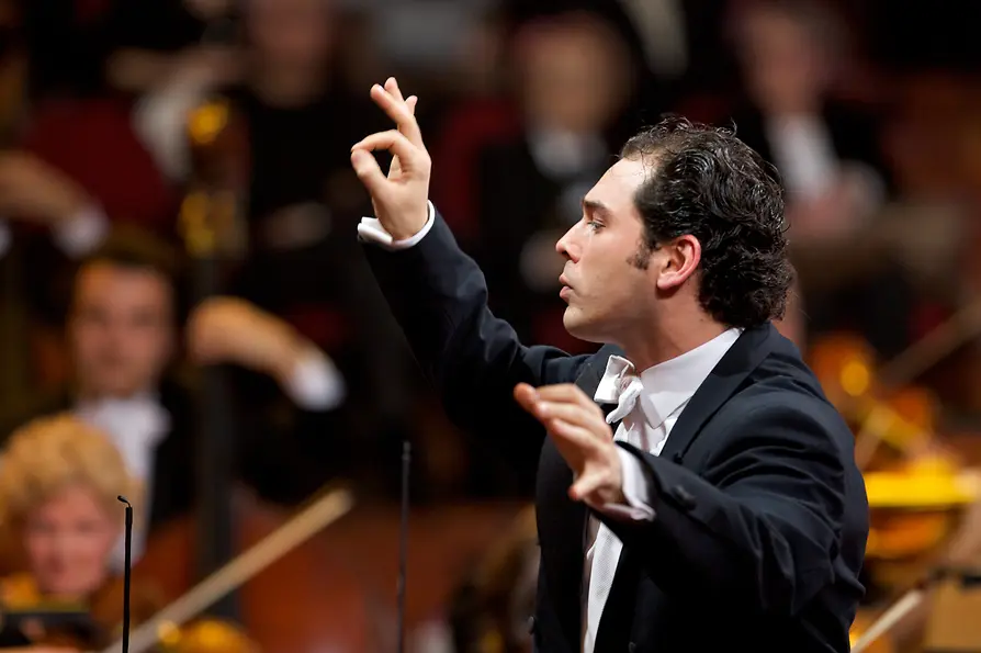 Conductor Tugan Sokhiev leads an orchestra on stage with raised hands.