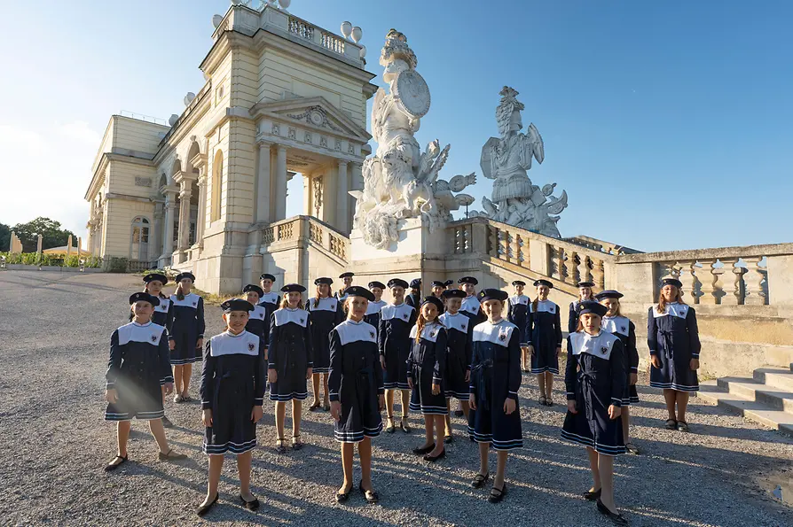 The Vienna Girls’ Choir stands in uniform in front of the Gloriette in Schönbrunn Palace Park, with historic architecture behind them.
