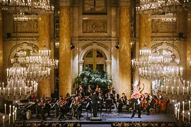 The Vienna Hofburg Orchestra performs in the ceremonial hall of the Hofburg, with marble columns and palm trees in the background, illuminated by large round chandeliers. 